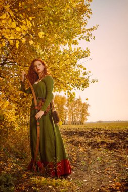 Tarihi yeniden yapılandırma. Muhteşem uzun kızıl saçlı, tarihi bir Kelt elbisesi olan güzel genç kadın okçu ormanın kenarında duruyor..