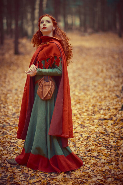 A beautiful red-haired girl of the Middle Ages poses looking up in the autumn forest. Celtic culture. Fantasy world.