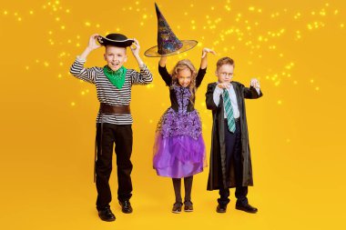 Cadılar Bayramı 'nda şeker mi şaka mı? Cadılar Bayramı kostümlü üç neşeli çocuk duygusal olarak stüdyoda sarı bir arka planda poz veriyor..