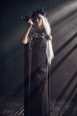 Güzel, görkemli siyah bir kraliçe, zengin başlıklı bir cadı ve zengin siyah bir elbise, karanlık bir kaledeki ışığın altında duruyor. Hayal Dünyası. 