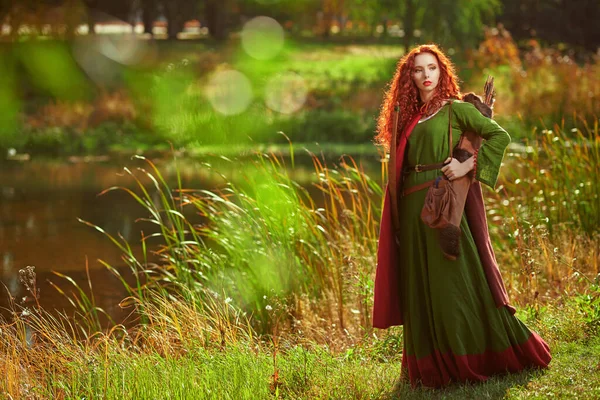Celtic culture. A beautiful red-haired girl archer of the Middle Ages stands next to the lake in sunny day. Fantasy world.