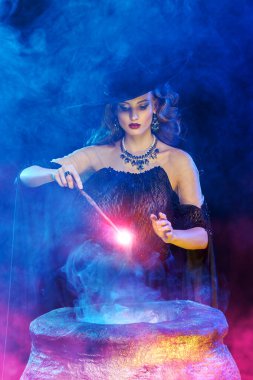 Cadılar Bayramı peri masalı. Şapkalı genç ve güzel bir cadı sihirli bir değnekle sihirli ateşlerle çevrili bir kazanı çağırır..