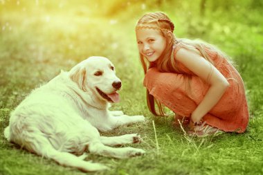 Parkta güneşli bir günde yeşil çimlerin üzerinde köpeğiyle oynayan hippi tarzı gülümseyen sevimli küçük kız. Yaz zamanı. Çocuklar ve evcil hayvanlar.