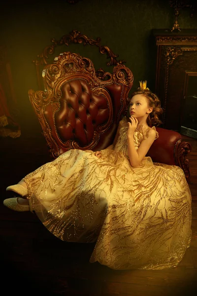 Fairy tales. Beautiful little princess girl in a golden dress and a crown sitting in an armchair in her vintage castle.