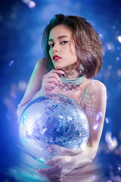 Beauty, Fashion. Glamorous young woman with evening make-up and hairstyle poses in jewelry with gems in the glow of disco ball. Party and holiday style.