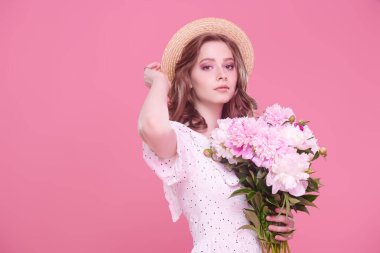 Açık beyaz yaz elbisesi ve hasır şapkalı, pembe arka planda şakayık demeti olan güzel bir kız. Yaz güzelliği ve moda. Mutlu insanlar..