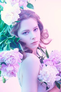 Güzellik, parfüm ve kozmetik. Soluk pembe şakayık çiçekleri arasında zarif leylak-menekşe makyajlı güzel bir genç kadının portresi.