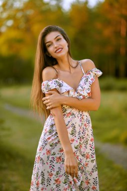 Romantik genç kadın parkta çiçekli bir elbiseyle poz veriyor ve gülümsüyor. Yaz modası ve güzelliği.