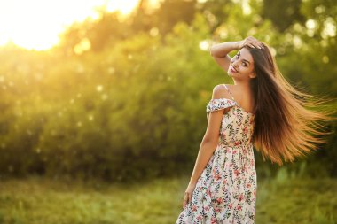 Mutlu genç kadın, parkta çiçekli bir elbiseyle hareket halindeyken poz veriyor. Yaz modası ve güzelliği.