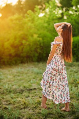 Güneşli bir günde çimenlikte çıplak ayakla çiçek desenli bir elbiseyle poz veren güzel bir genç kızın tam boy portresi. Yaz modası ve güzelliği.