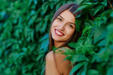 Çekici kahverengi saçlı genç kadının portresi parktaki yeşil bir çitin arka planına gülümsüyor. Yaz modası ve güzelliği.