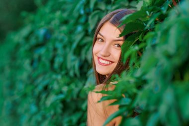 Yaz modası ve güzellik. Çekici kahverengi saçlı genç kadın portresi parktaki yeşil bir çitin arka planına gülümsüyor..