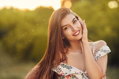 Romantik genç bir kadın yaz çiçekli bir elbiseyle poz veriyor ve parkta gülümsüyor. Yaz modası ve güzelliği.