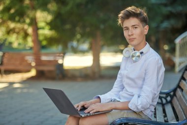 Bir yaz parkında dizüstü bilgisayar kullanan ve gülümseyen mutlu bir gencin portresi. Güneşli bir yaz günü. Eğitim.