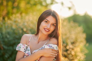 Mutlu yaz zamanları. Gün batımında parlak çiçekli bir elbise giymiş çekici bir kızın portresi. Yaz modası ve güzelliği.