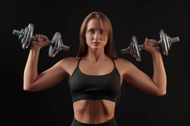 Güzel atletik genç kadın siyah spor kıyafetleriyle poz veriyor. Kollarında halterler var. Siyah bir arkaplanda stüdyo resmi.