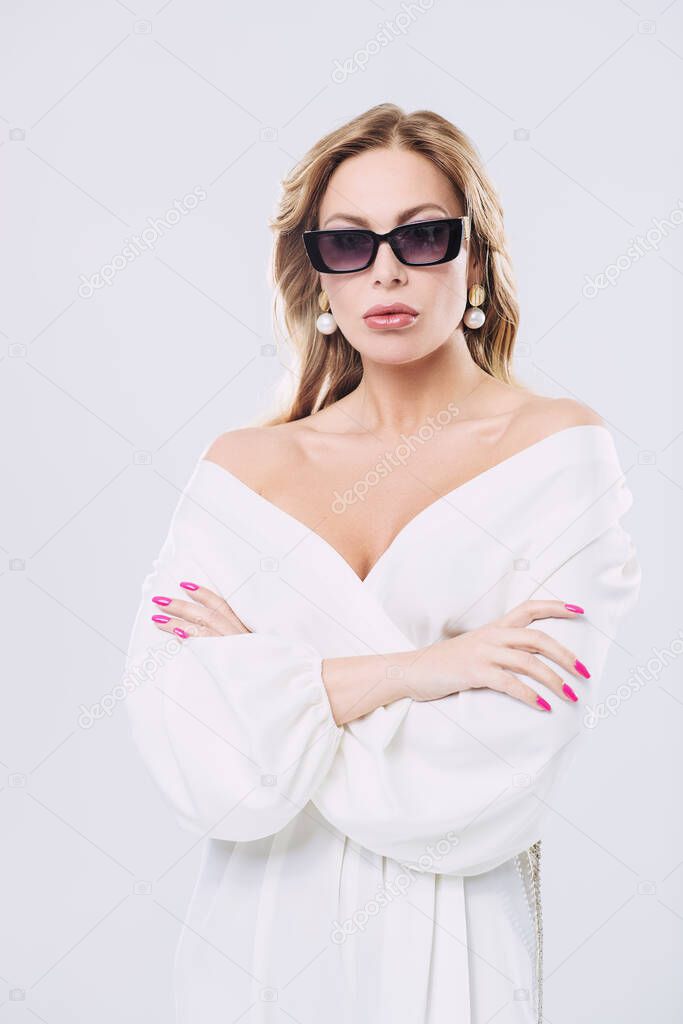 Portrait of a stylish sexy middle aged woman in a white suit, precious jewelry and sunglasses on a white background. Beauty, fashion. 
