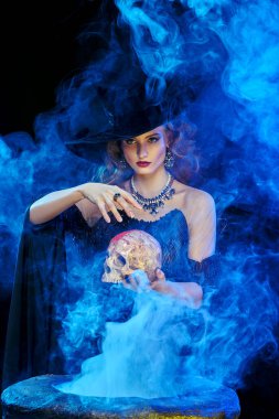 Cadılar Bayramı büyüsü. Şapkalı ve zarif giysili güzel bir cadı sihirli dumanla çevrili bir kazanın üzerinde kafatasıyla büyüler..