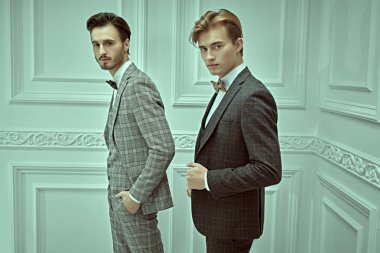 Men's style. Portrait of two elegant young men in classic three-piece suits standing in white luxury apartment. clipart