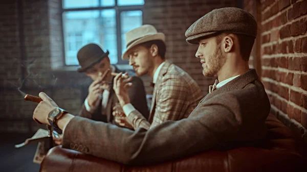 Three handsome men in elegant suits sit on a leather sofa, smoke cigars and read newspapers. Newspaper editorial office. Retro style. Men\'s fashion.