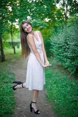 Uzun saçlı, parkta yürüyen ve mutlu bir şekilde gülümseyen romantik bir genç kadının tam boy portresi. Yaz modası ve güzelliği.