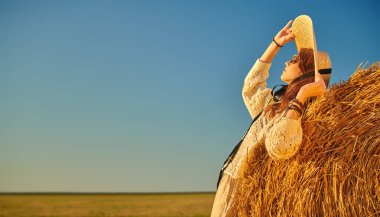 Örgü kazaklı, geniş kenarlı hasır şapkalı ve yuvarlak gözlüklü zarif genç bir kadın samanlıkta samanlıkta gün batımını seyrediyor. Yaz ve sonbahar modası. Boşluğu kopyala. 