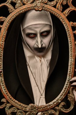 Halloween. Portrait of a scary devilish nun looking through a vintage picture frame on a black background. Horrors. clipart