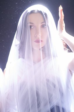 Kafasında beyaz bir peçeyle siyah bir arka planda ışıkla çevrili güzel esmer bir kızın portresi. Güzel sanatlar, moda. Stüdyo resmi.