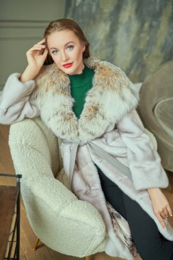 Kürk manto modası. Pahalı vizon kürklü ve vaşak kürklü güzel sarışın kadın beyaz koltukta poz veriyor. Klasik lüks bir iç mekanda portre. 