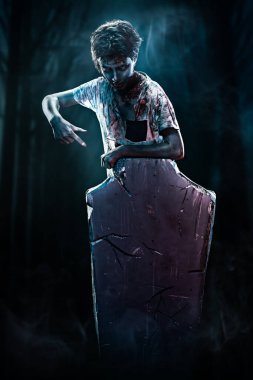 Korkunç Cadılar Bayramı. Korkunç zombi çocuk mezar taşının arkasında duruyor ve yüzeyindeki fotokopi alanını gösteriyor. Ürkütücü arkaplan.