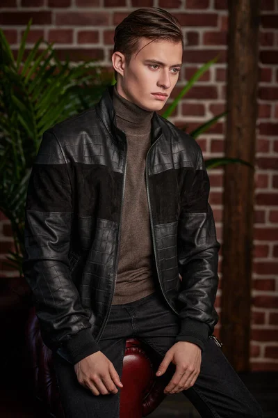 Attractive young man in a leather jacket leaning on a red sofa is posing in the background of a brick wall. Portrait in a loft interior design. Men\'s fashion. Lifestyle.