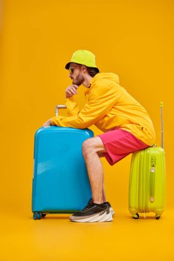 Parlak şık spor kıyafetleri içinde havalı genç bir adam bavullarıyla oturur ve tatilinin başlamasını bekler. Tatil ve seyahat. Sarı arka planda tam boy stüdyo resmi.