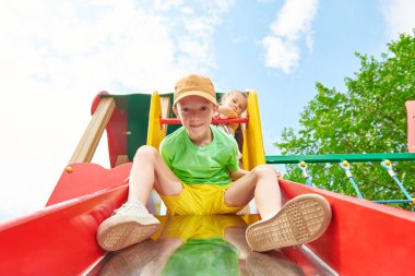 Gülümseyen çocuk oyun parkında kaydırağın üzerinden neşeyle iniyor. Gökyüzü ve ağaçların arka planında tam boy portre. Mutlu bir çocukluk. Çocuk oyunları ve yaz tatilleri. 
