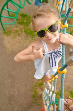 Güneş gözlüklü mutlu küçük kız oyun parkında iple tırmanırken eğleniyor. Çocuk oyunları ve yaz tatilleri. Mutlu çocukluk.