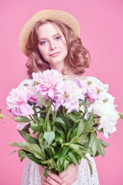 Açık beyaz bir yaz elbisesi ve elinde şakayık buketi tutan hasır şapkalı güzel bir kız. Pembe arka planda stüdyo portresi. Yaz güzelliği ve moda. Mutlu insanlar..