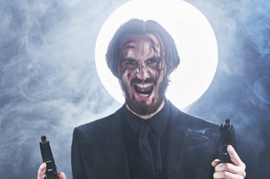 Siyah takım elbiseli, elinde silahlarla ölümcül bir savaşa hazır agresif bir aksiyon kahramanı. Karanlık sisli arka plan, arkasında ışık var..