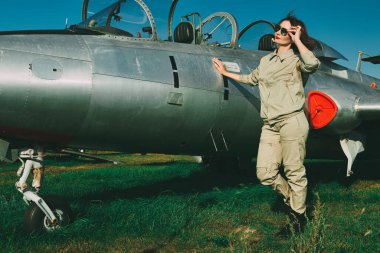 Full length portrait of a beautiful woman pilot wearing uniform and sunglasses posing at her fighter jet. Military and civil aircraft.  clipart