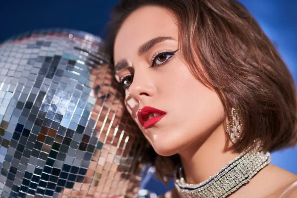 Party and holiday style. Close-up portrait of a glamorous young woman with elegant evening makeup and hairstyle holding a disco ball next to her face. Beauty, Fashion and Jewelry.