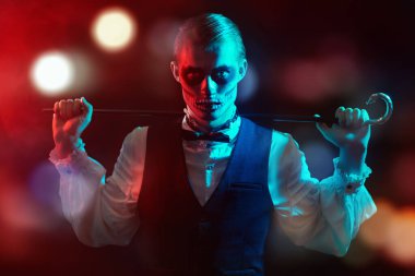 Kafatası makyajı ve bastonla poz veren şık, zarif bir adam. Karnaval gece lambaları olan karanlık bir arka plan. Dia de los muertos. Ölüler Günü. Cadılar Bayramı.