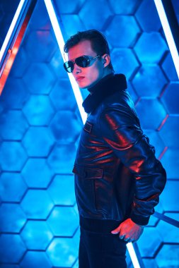 Siyah güneş gözlüklü ve siyah deri ceketli yakışıklı adam neon lambaların arasında duruyor. Erkek tarzı ve modası. Futurizm, tekno stil.