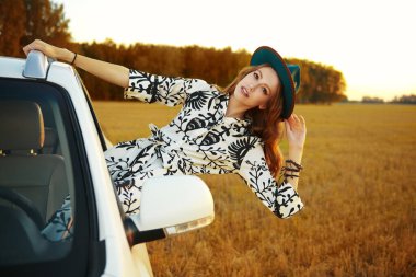 Göz kamaştırıcı giyinmiş, boho tarzı pozlar veren genç bir kadın arabanın camından sarkıyor. Moda, sonbahar havası. Kırsal arka planda batan güneşin ışınlarında.