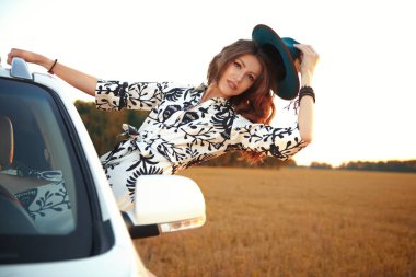 Boho tarzı elbiseli ve şapkalı zarif genç bir kadın arabanın camından neşeyle sarkıyor. Sonbahar tatili. Moda, sonbahar havası..