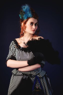 Viktorya dönemi tarzı makyaj ve saç stili olan çekici kızıl saçlı bir kadının portresi, 19. yüzyıl modasında bir hayranla poz veriyor. Koyu mavi arkaplan. 