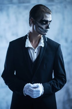 Kafatası makyajlı, zarif siyah takım elbiseli ve grunge arka planda beyaz gömlekli yakışıklı bir genç. Dia de los muertos. Ölüler Günü. Cadılar Bayramı.