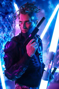 Koruyucu kostümlü cesur siber punk savaşçısı elinde silahla neon lambaların arasında duruyor. Oyun, sanal gerçeklik. 
