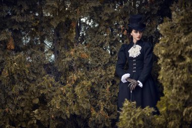 19. yüzyılın sonları ve 20. yüzyılın başlarına ait tarihi yeniden yapılanma. Katı siyah elbiseli ve şapkalı zarif, koyu renk saçlı bir bayan parkta yürüyor. Tarihsel makyaj ve saç stili.