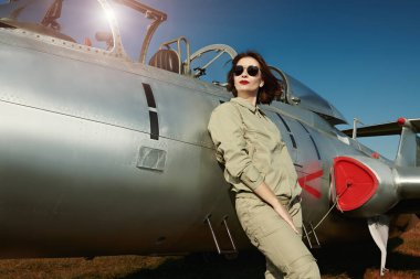 Military and commercial aircraft. Portrait of a confident pilot woman in uniform and sunglasses posing next to her fighter jet.  clipart