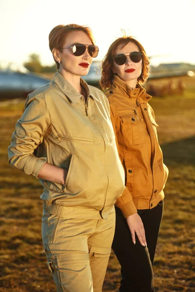 Portrait of two gorgeous pilots women in stylish uniform and sunglasses posing on the airfield. Commercial aviation.