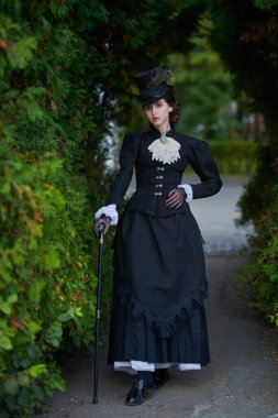 19. yüzyılın sonları ve 20. yüzyılın başlarına ait tarihi yeniden yapılanma. Katı siyah elbiseli ve şapkalı zarif, koyu renk saçlı bir bayan parkta yürüyor. Tarihsel makyaj ve saç stili.