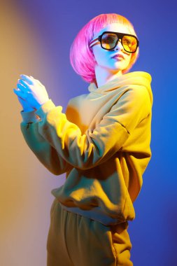 Gençlik modası ve güzellik trendleri. Kendine güvenen, pembe saçlı, sarı güneş gözlüklü, sarı kapüşonlu ve karışık renkli ışıkta duran modern bir kız. Spor şıklığı modası. 